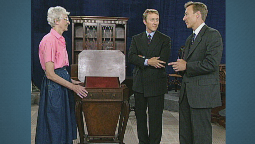 An older woman, Leigh Keno, stands to the left of a Duncan Phyfe sewing table, ca. 1805 and appraises its value as two men in suits stand on the right, discussing.