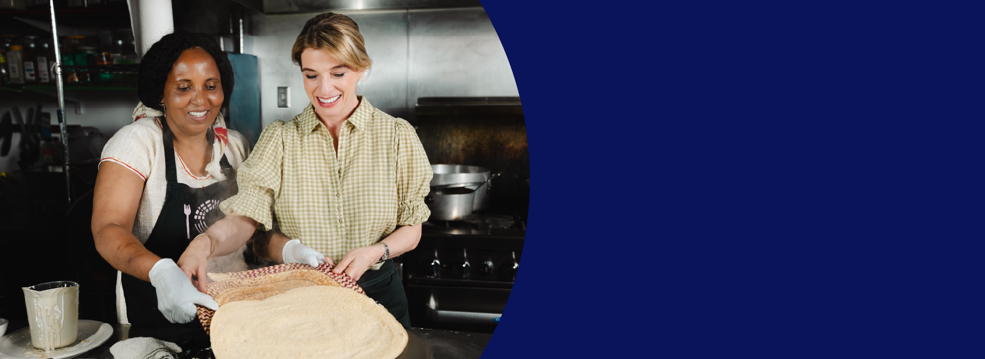 Two women stand next to each other in the kitchen. One demonstrates to the other how to handle a large circular dough.