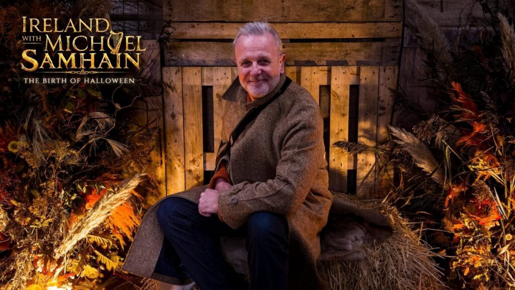 Michael Londra, a middle aged man, sits on a straw palette while wearing a long, brown, tweed coat. He sits in front of a wooden fence and is surrounded by purposefully staged fall foliage and decor.