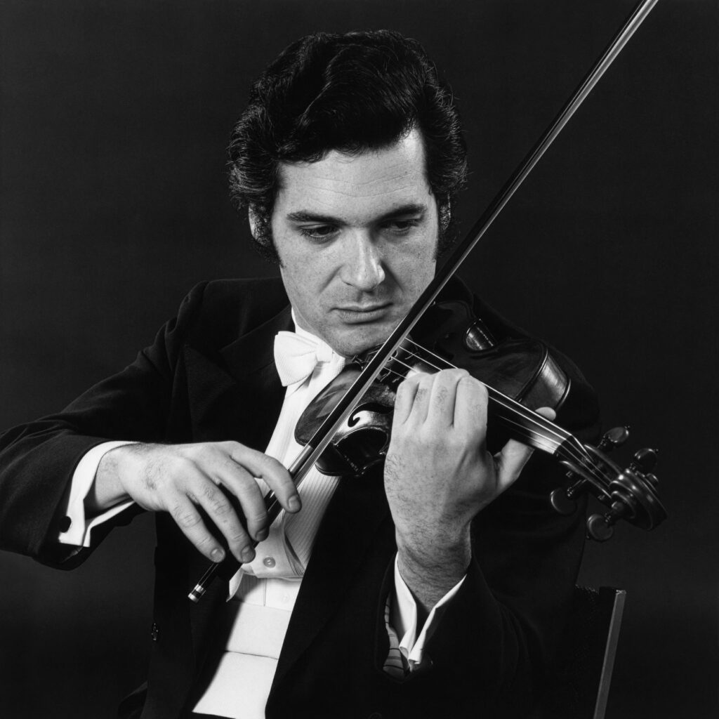 In this black and white photo a man, Pinchas Zukerman, wears a black tuxedo and plays the violin.