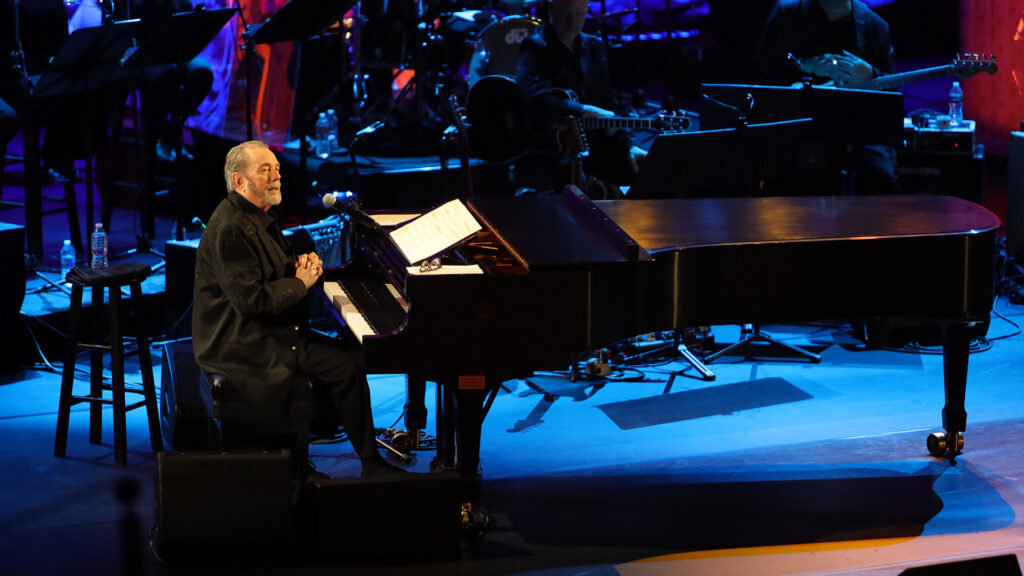 An older man, which can be identified as iconic American songwriter Jimmy Webb, sits at a grand piano, surrounded by a symphony on stage. The blue stage lights cast dramatic shadows on the stage.
