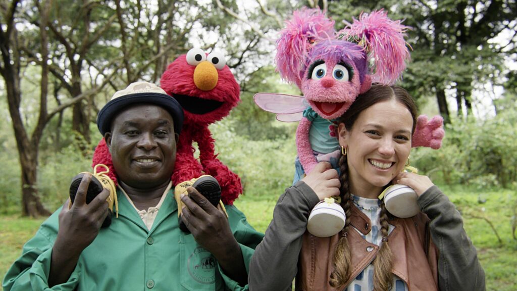 Two Sesame Street characters, Elmo and Abby Cadabby, sit on the shoulders of two adults. The are are all outside and smiling.