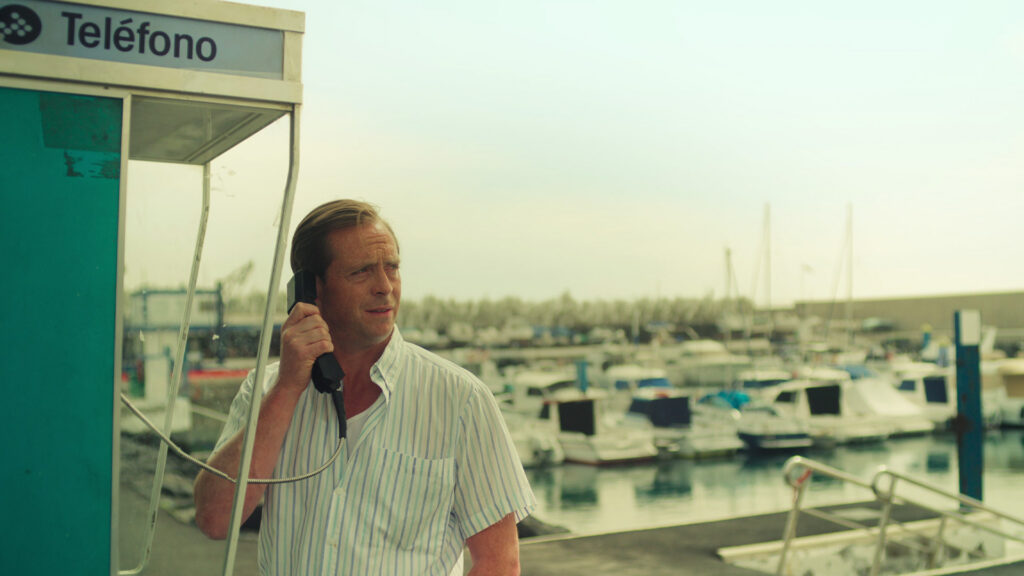 A white middle aged man uses a payphone while standing on a boat dock. He looks into the distance with a confused expression.